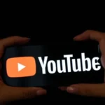 YouTube logo displayed on a smartphone illustrating the platform's trial to reduce its minimum creator payment threshold from $100 to $20.